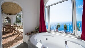 Un baño con una bañera grande, vistas al mar a través de una ventana y un balcón con plantas en macetas.