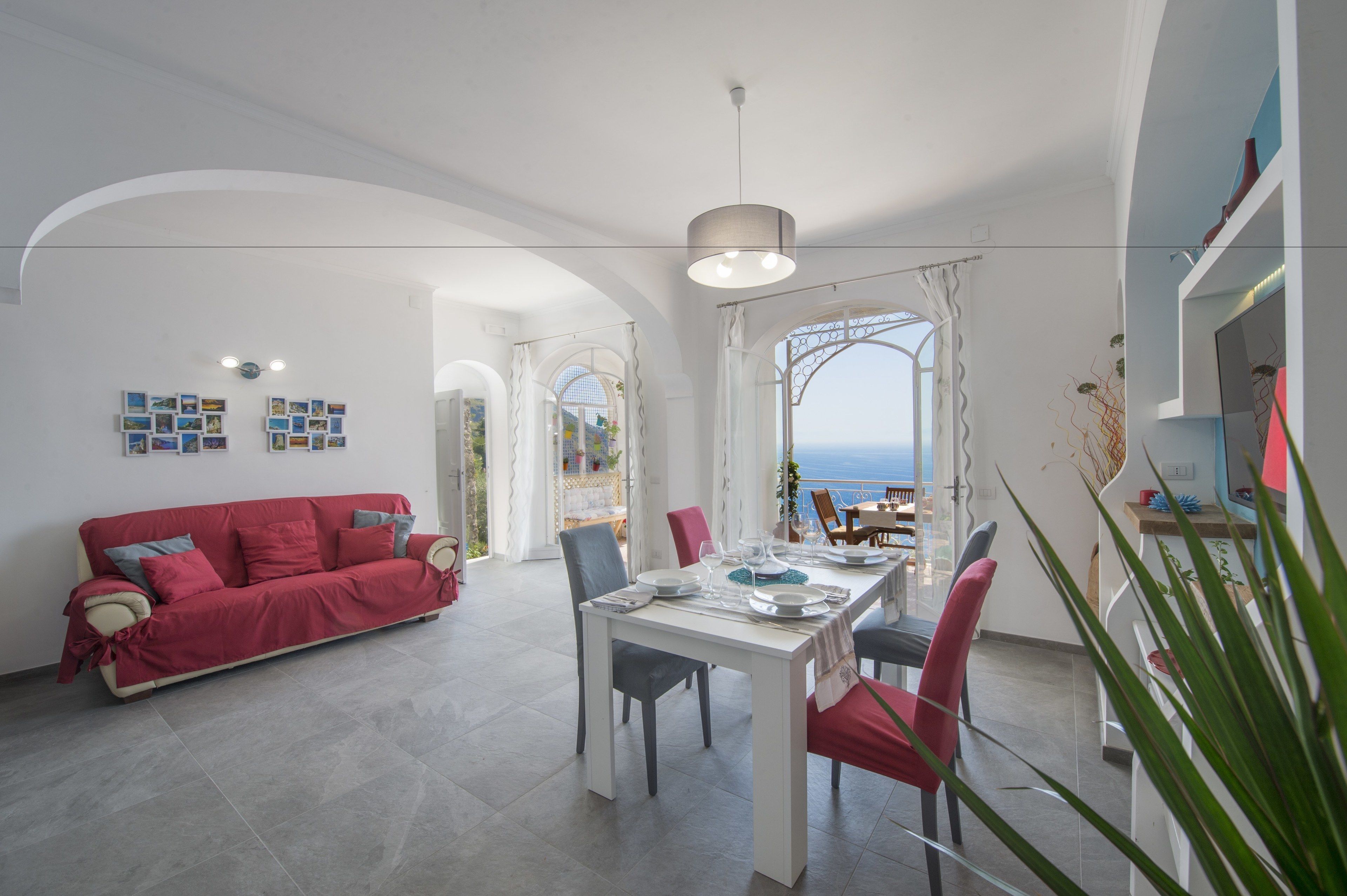 A bright living room with a red sofa, a dining table set for two, and a view of the sea through a large window.