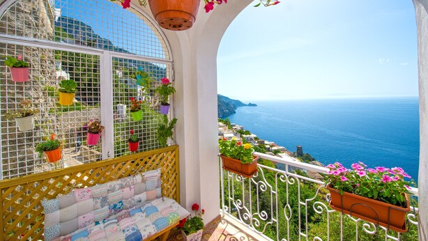 Un balcón con vista al mar, plantas en macetas y una zona de estar con cojines.