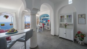 Amplia habitación con balcón vistas al mar, que incluye una zona de comedor con mesa y sillas, un área de cocina con armarios y un televisor, y una sala de estar con sofá y mesa de centro.