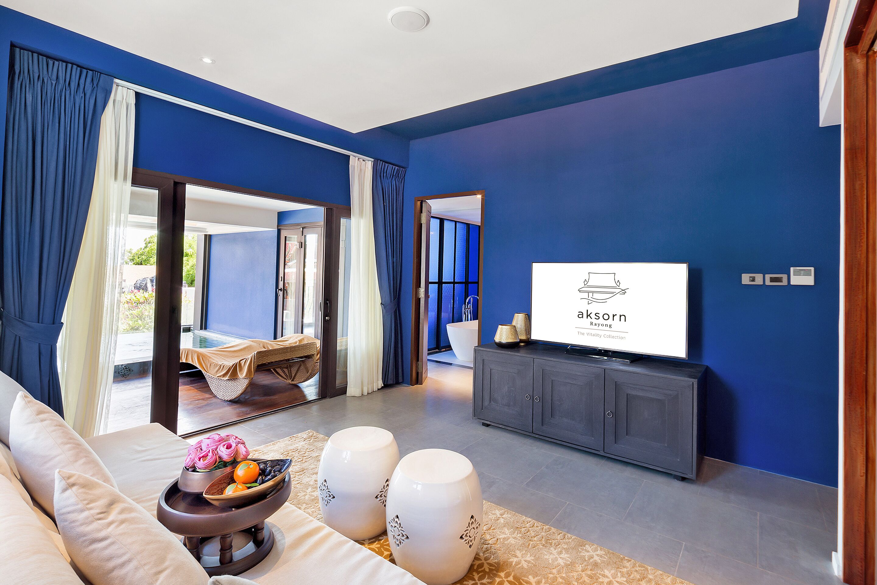 A modern living room with a sofa, a coffee table with decorative items, a flat-screen TV on a cabinet, and a view of a pool area through glass doors.