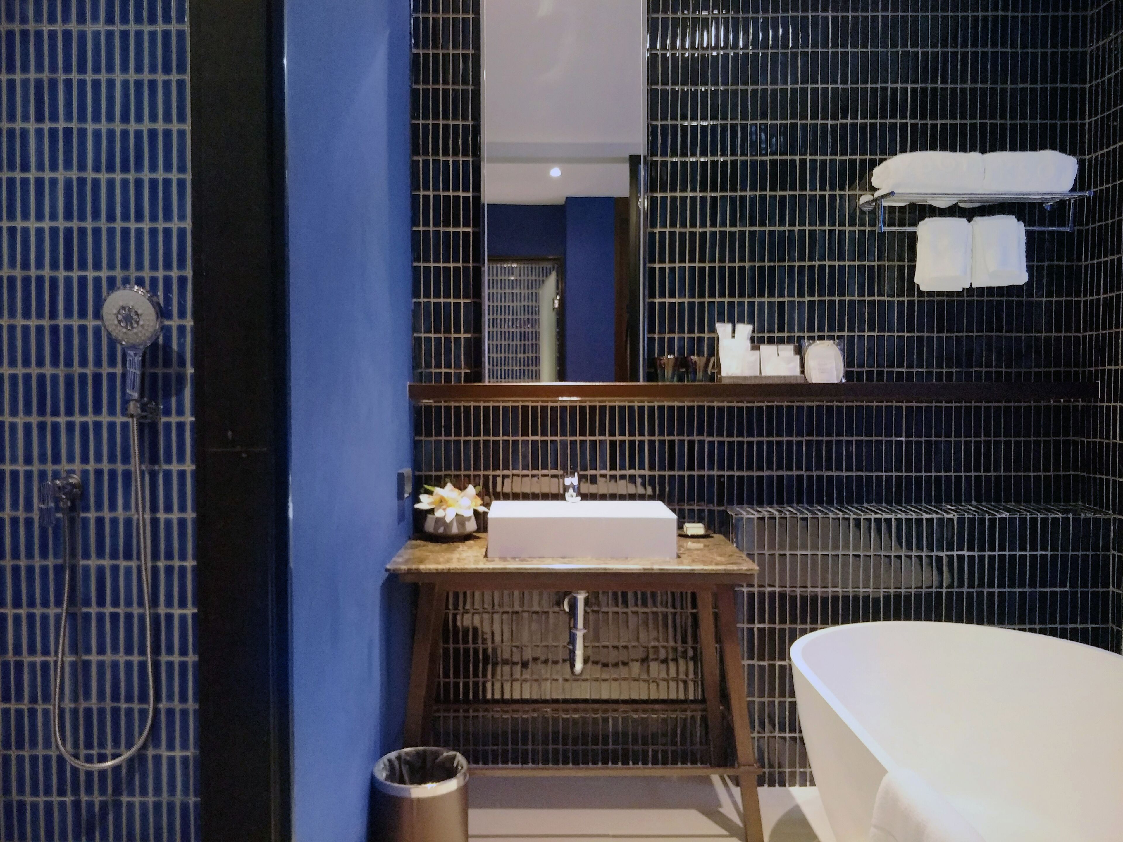 A modern bathroom with a blue tiled wall, a wooden vanity with a sink, a mirror, and a white bathtub.
