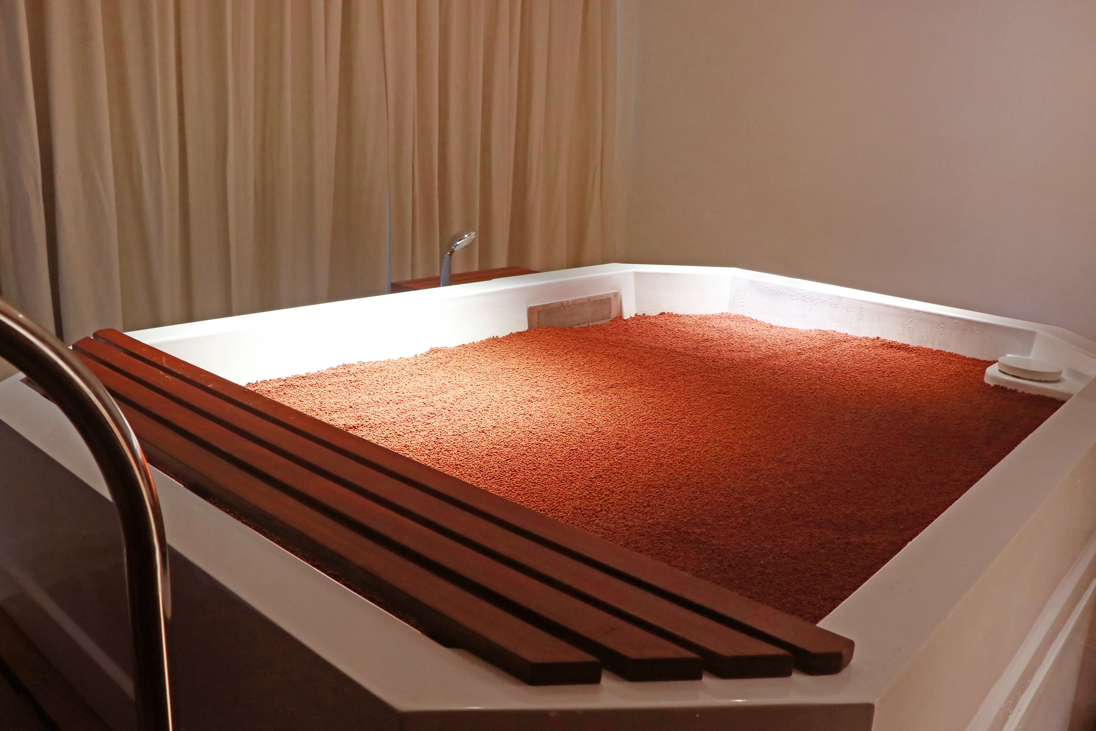 A bathtub filled with reddish-brown bath salts, a wooden bench, and a showerhead.