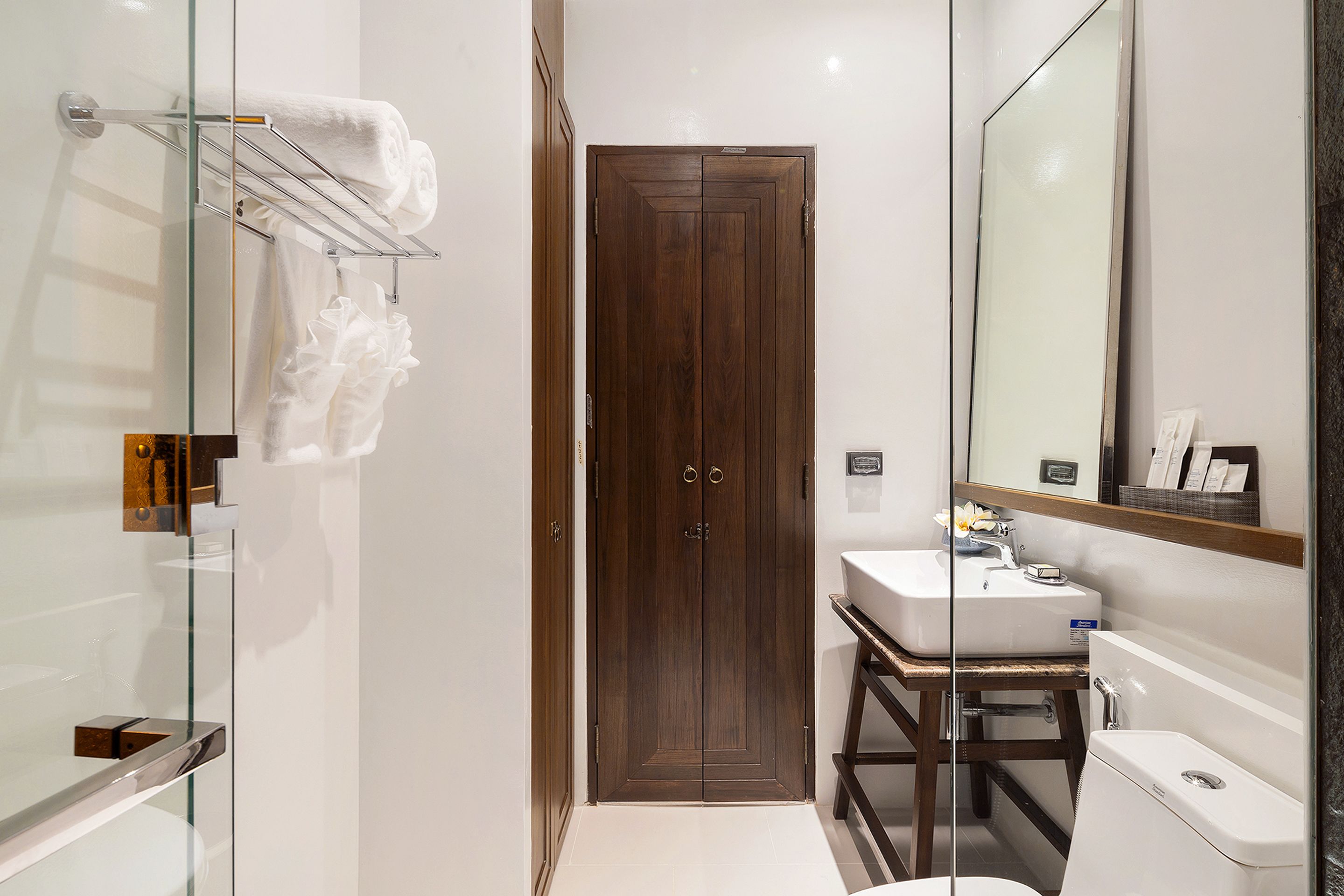 A modern bathroom with a glass shower enclosure, a wooden door, a white sink, and a large mirror.
