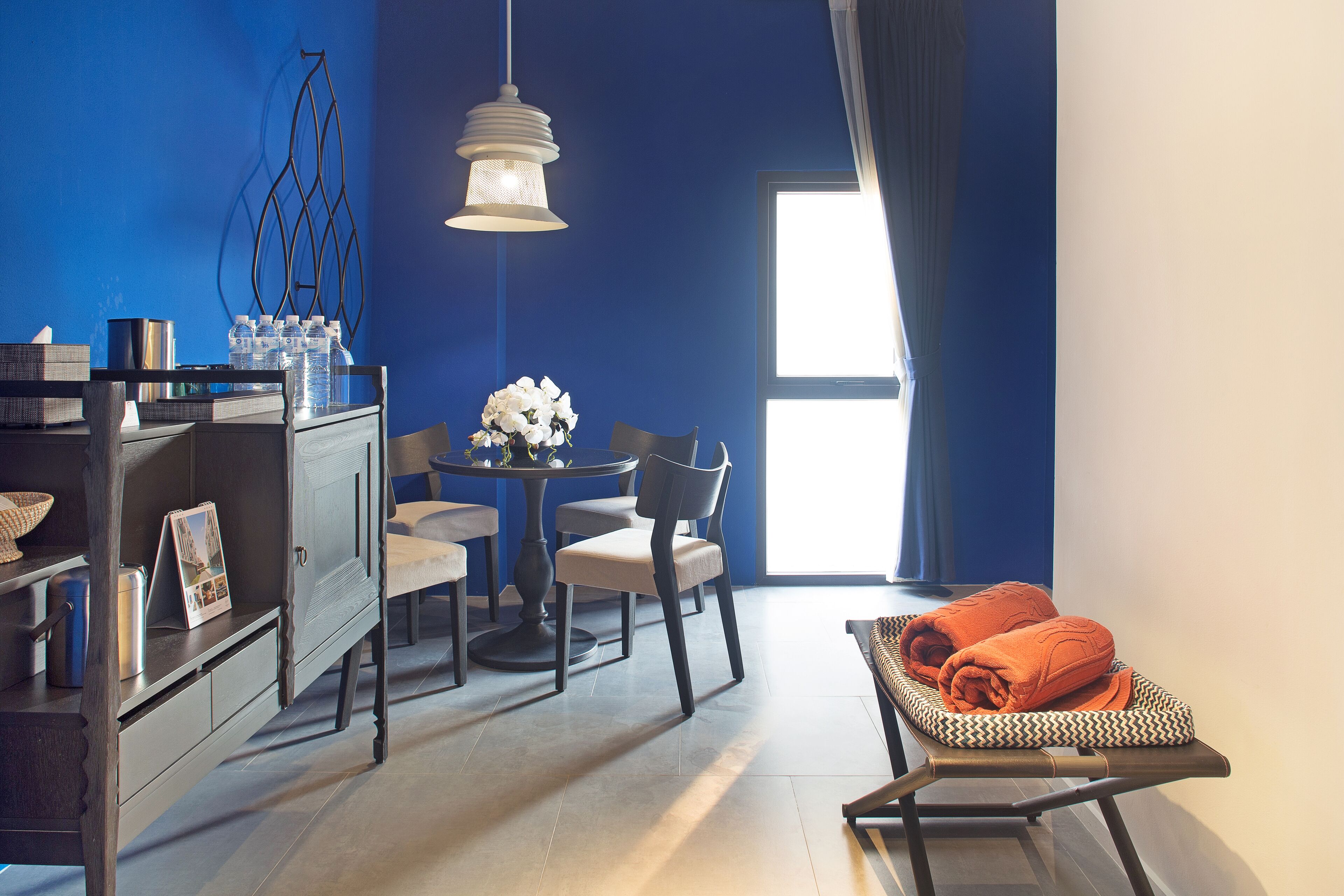 A room with a blue wall, a dining table with chairs, a small cabinet, a hanging light fixture, and a bed with an orange blanket.