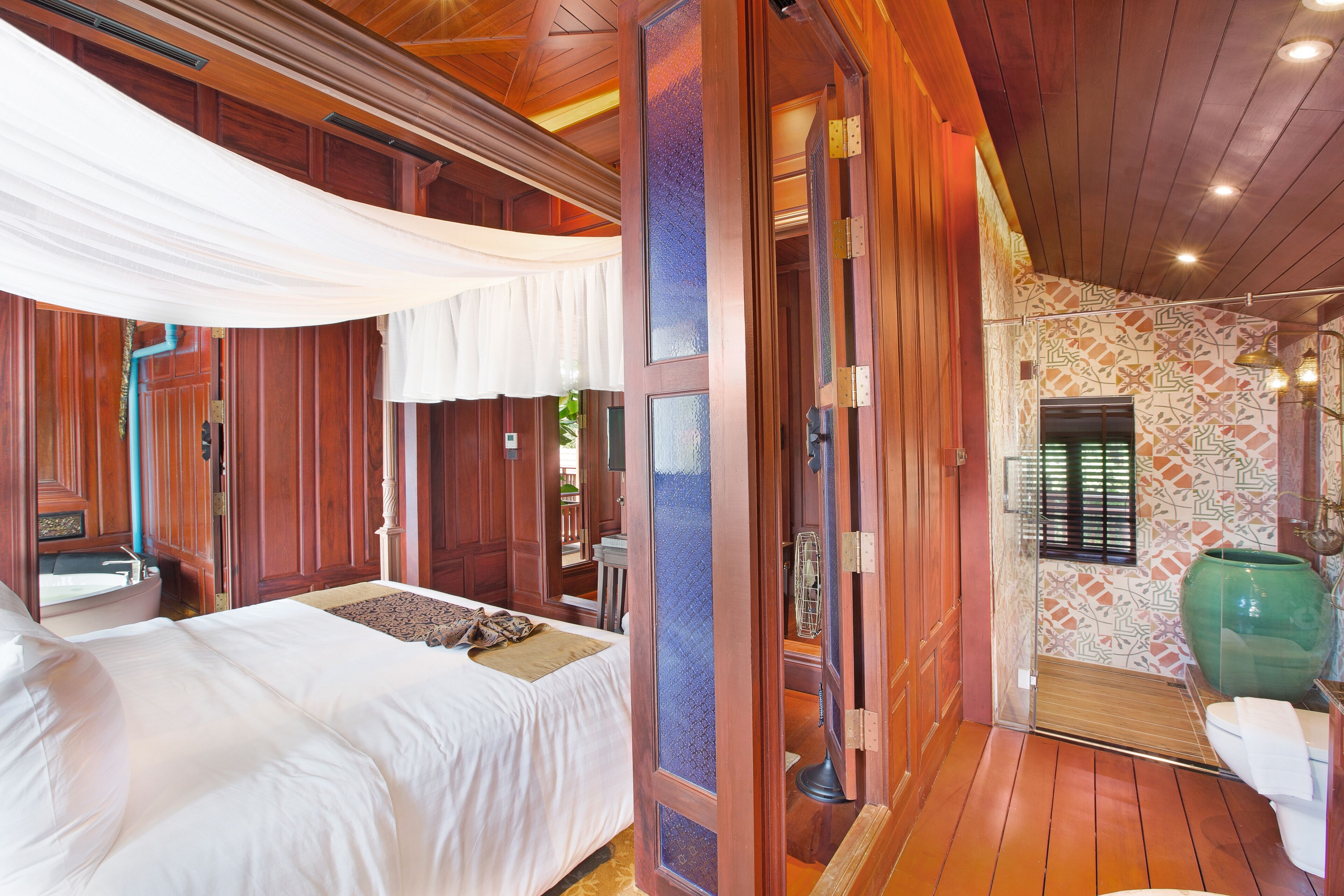 Una habitación con piso de madera, una cama con ropa blanca, una puerta de madera y un baño con pared estampada y lavamanos verde.