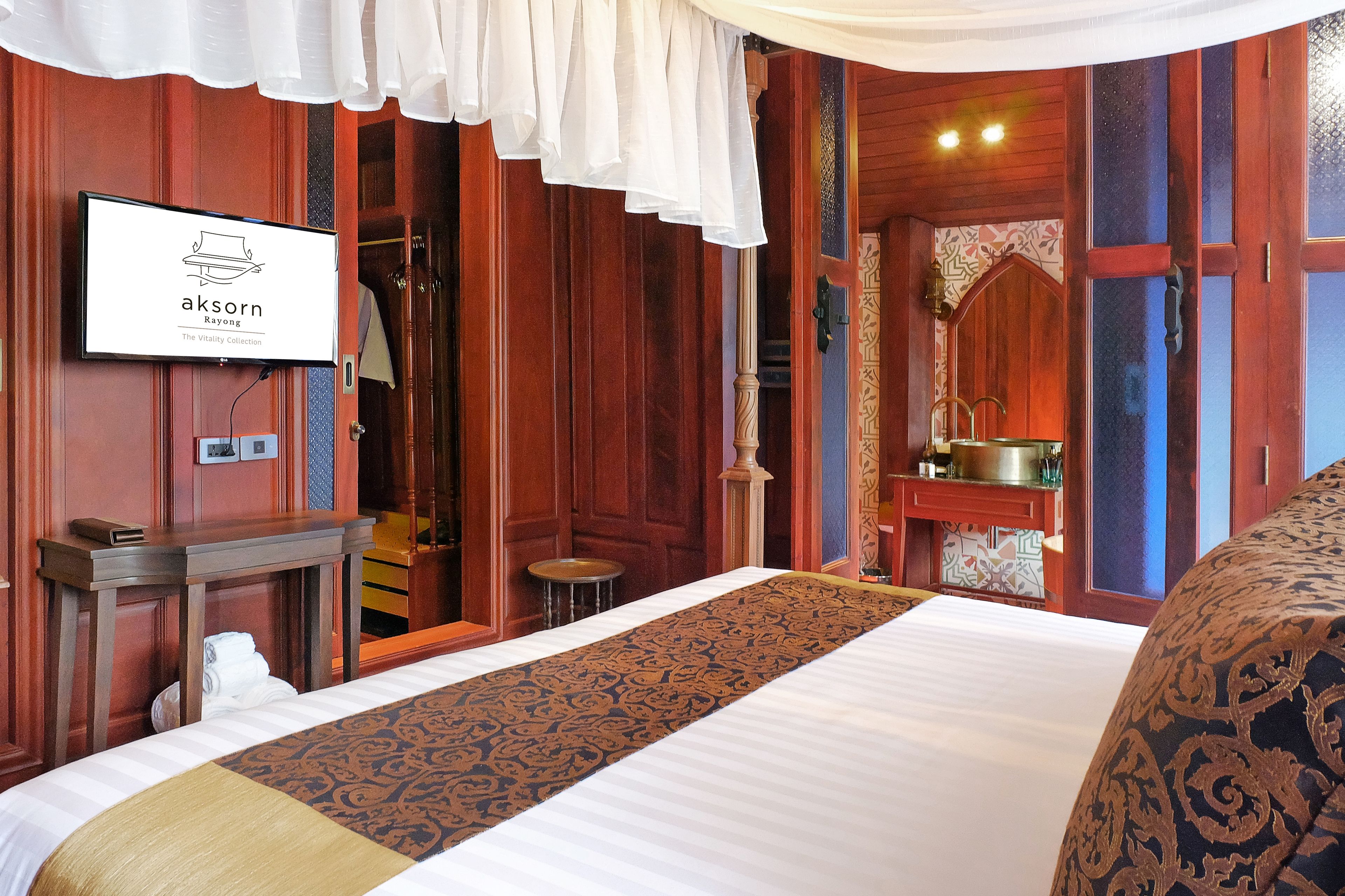 Quarto de hotel com uma cama, televisão exibindo 'akorn Rayong', uma escrivaninha de madeira, um banquinho pequeno e um banheiro com espelho e pia.