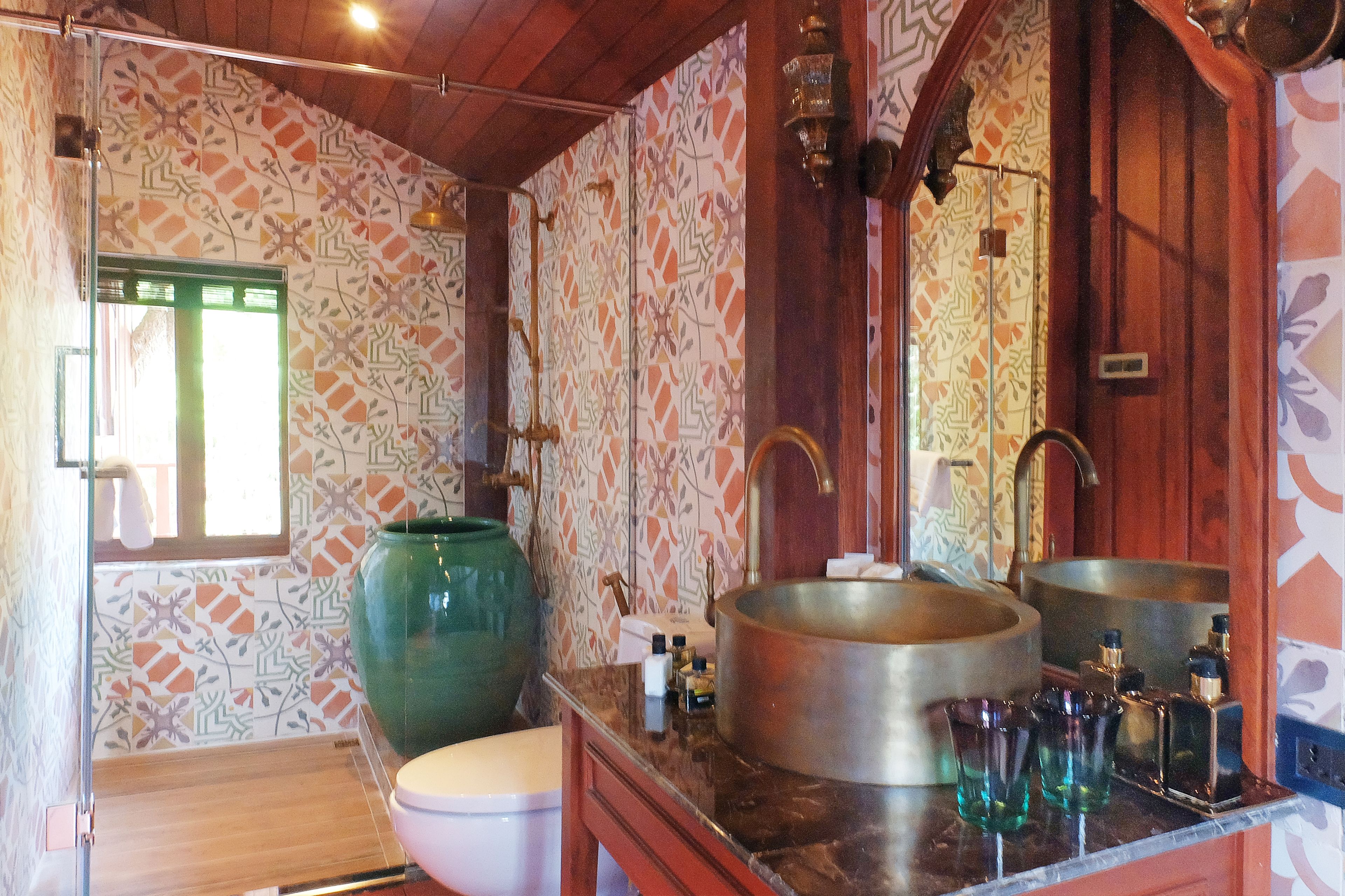 A bathroom with patterned tiles, a large green vase, a sink with two basins, and a mirror.