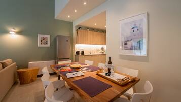 Una sala de estar moderna con una mesa de comedor de madera, sillas blancas, una pequeña mesita auxiliar de madera y un sofá. Se ve una cocina al fondo con armarios de madera y una encimera blanca.
