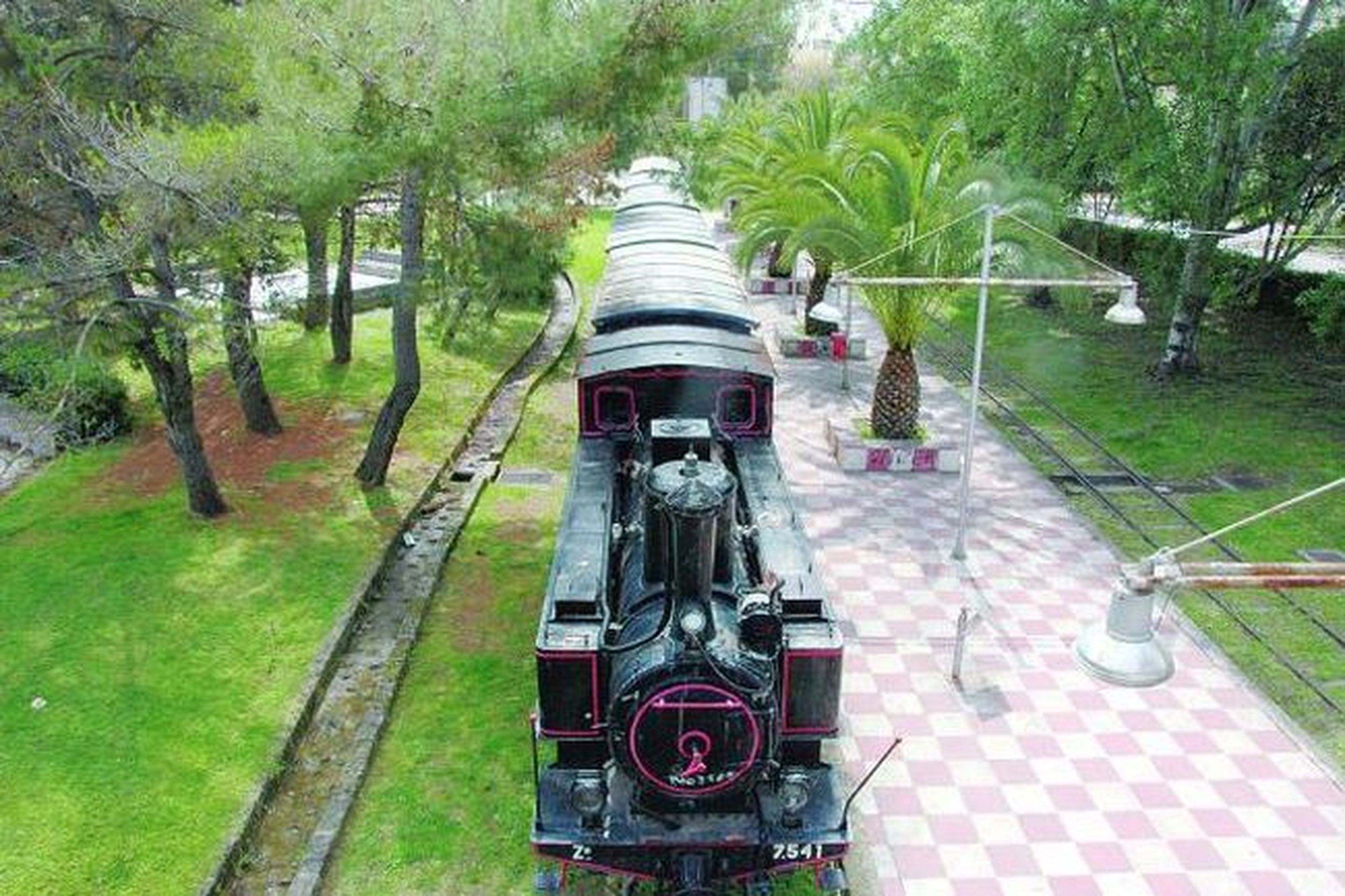 Una locomotora de vapor antigua sobre rieles, rodeada de follaje y un pavimento a cuadros.