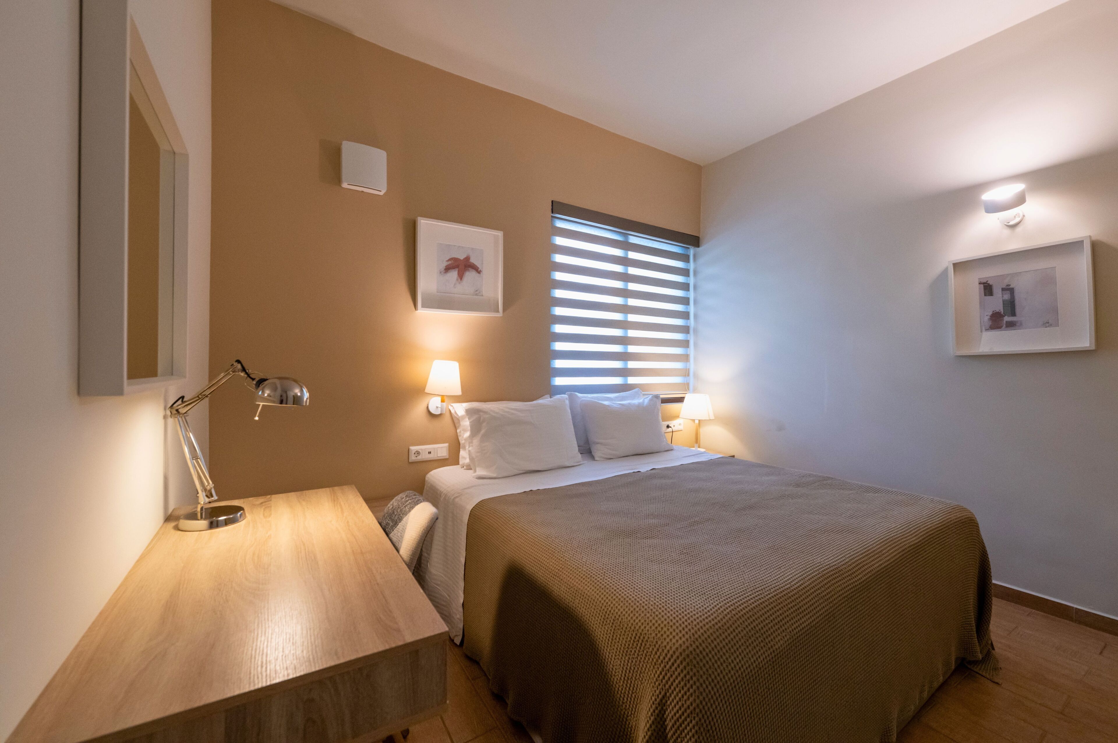 A bedroom with a bed, bedside lamps, a nightstand, a window with blinds, and wall-mounted artwork.