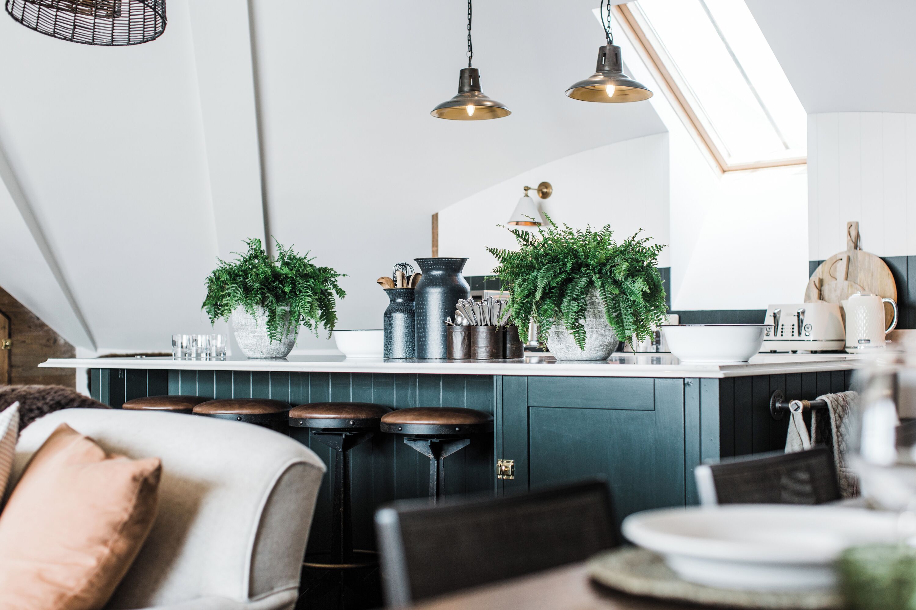 A modern kitchen with dark cabinetry, a central island with stools, pendant lighting, and potted plants.