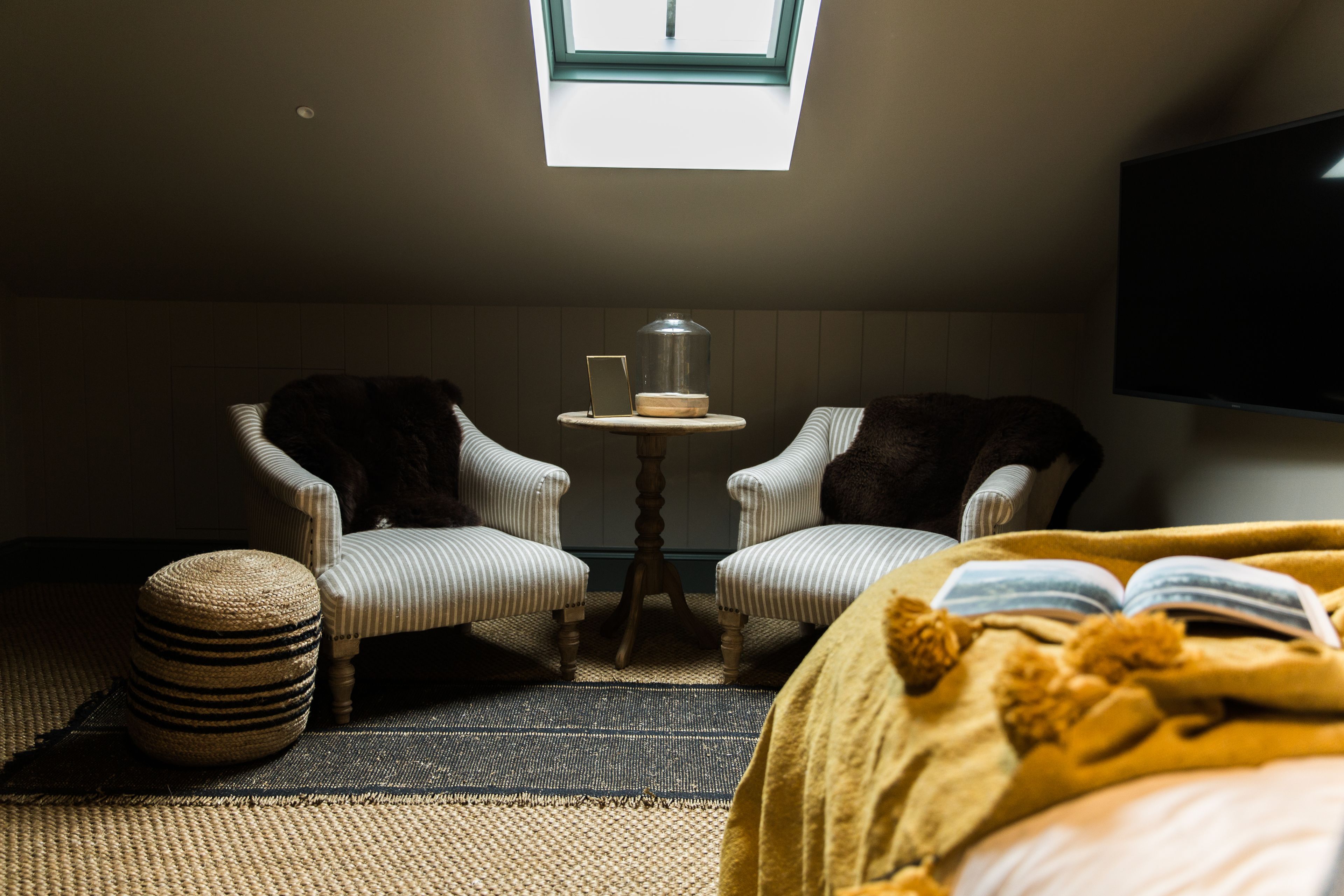 A cozy room with two upholstered chairs, a small round table with a glass dome, a large bed with a yellow blanket, and a television mounted on the wall.