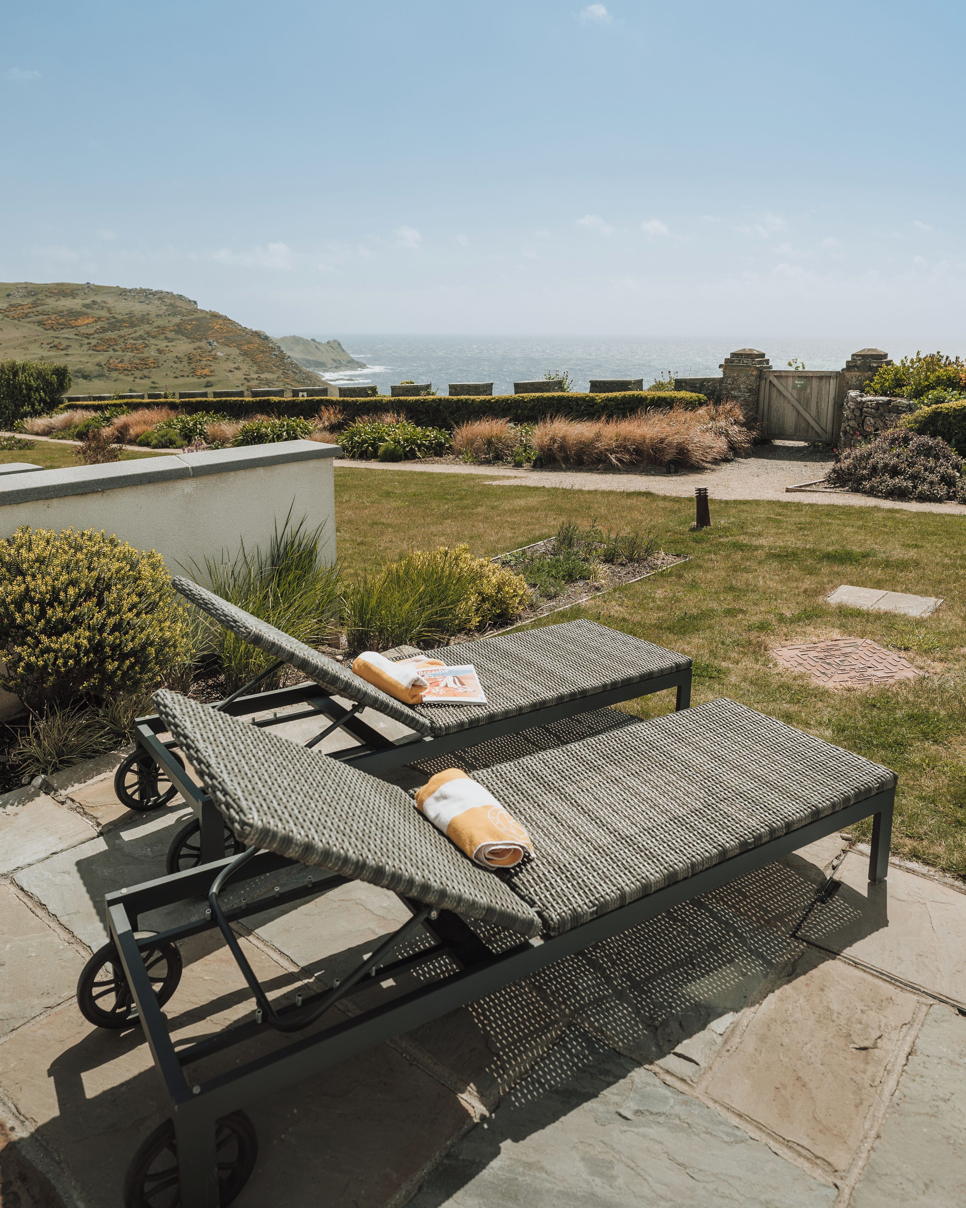 A patio with two lounge chairs and a table, overlooking a scenic view of the ocean and hills.