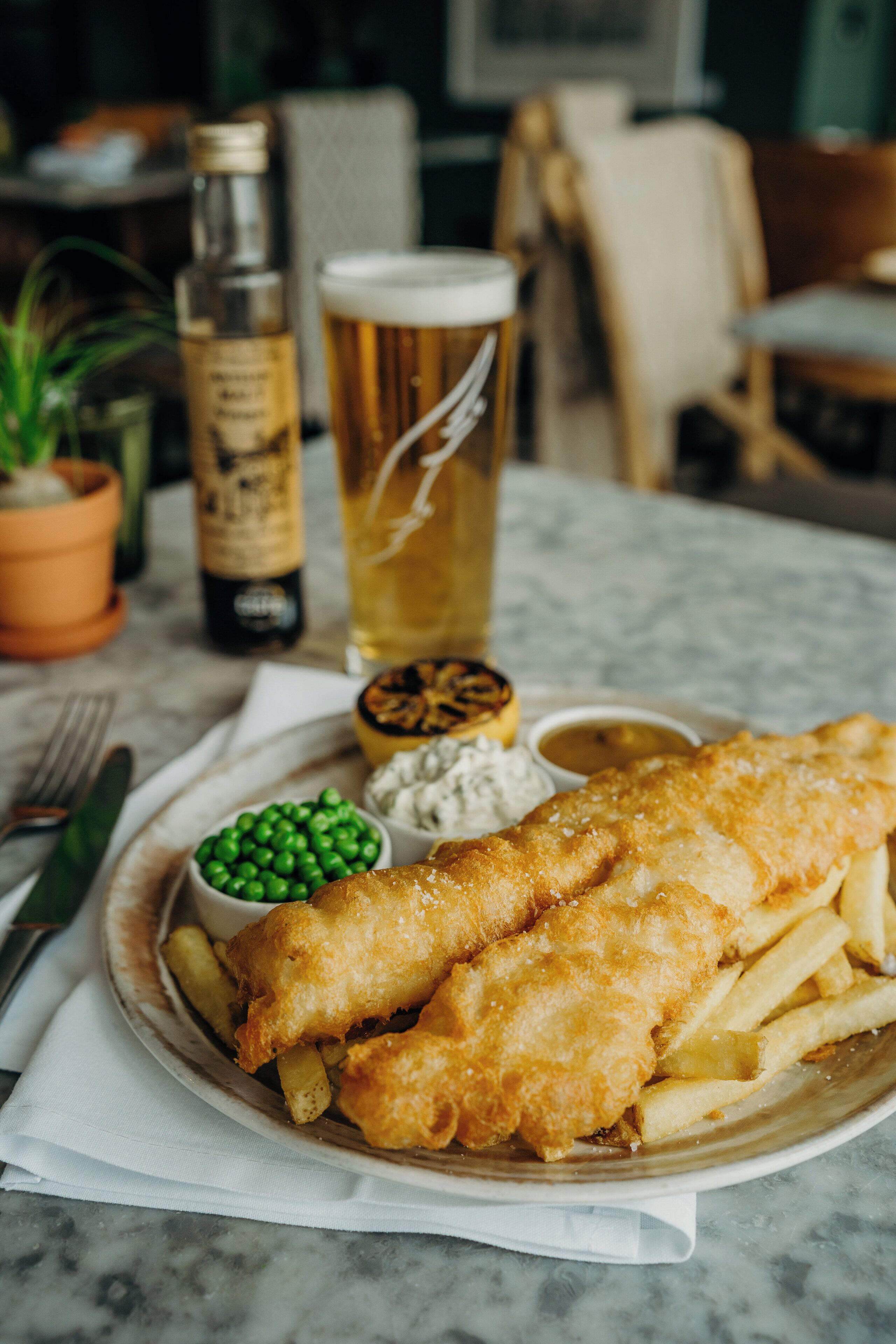 Un plato de pescado con patatas fritas, una jarra de cerveza, una botella y un cuenco pequeño de guisantes sobre una mesa.