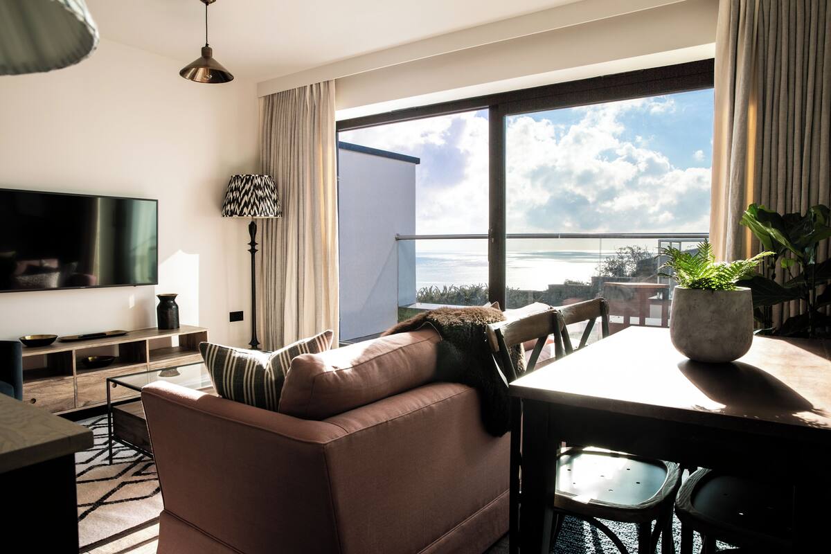 A modern living room with a sofa, TV, dining table, and a view of the sea.