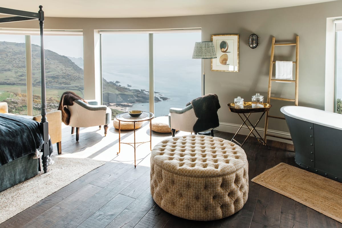 A bedroom with a large window overlooking a scenic landscape, a bed, a chair, a round ottoman, a small table with decorative items, and a bathtub.