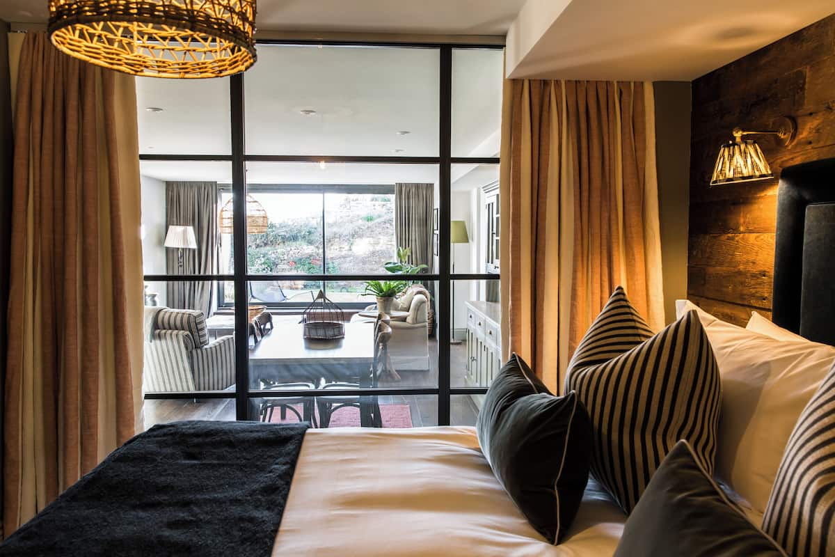 A modern hotel room with a large window, a bed with striped pillows, a black throw, and a woven light fixture.