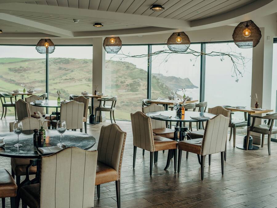 Un espace repas avec des tables et des chaises rondes, de grandes fenêtres offrant une vue sur les collines et des lampes suspendues.