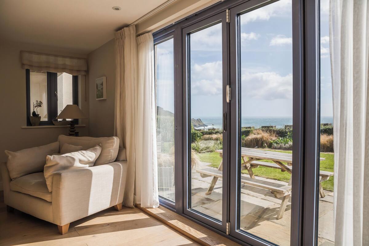 A room with a sofa, a lamp, and a view of a garden and beach through large sliding glass doors.