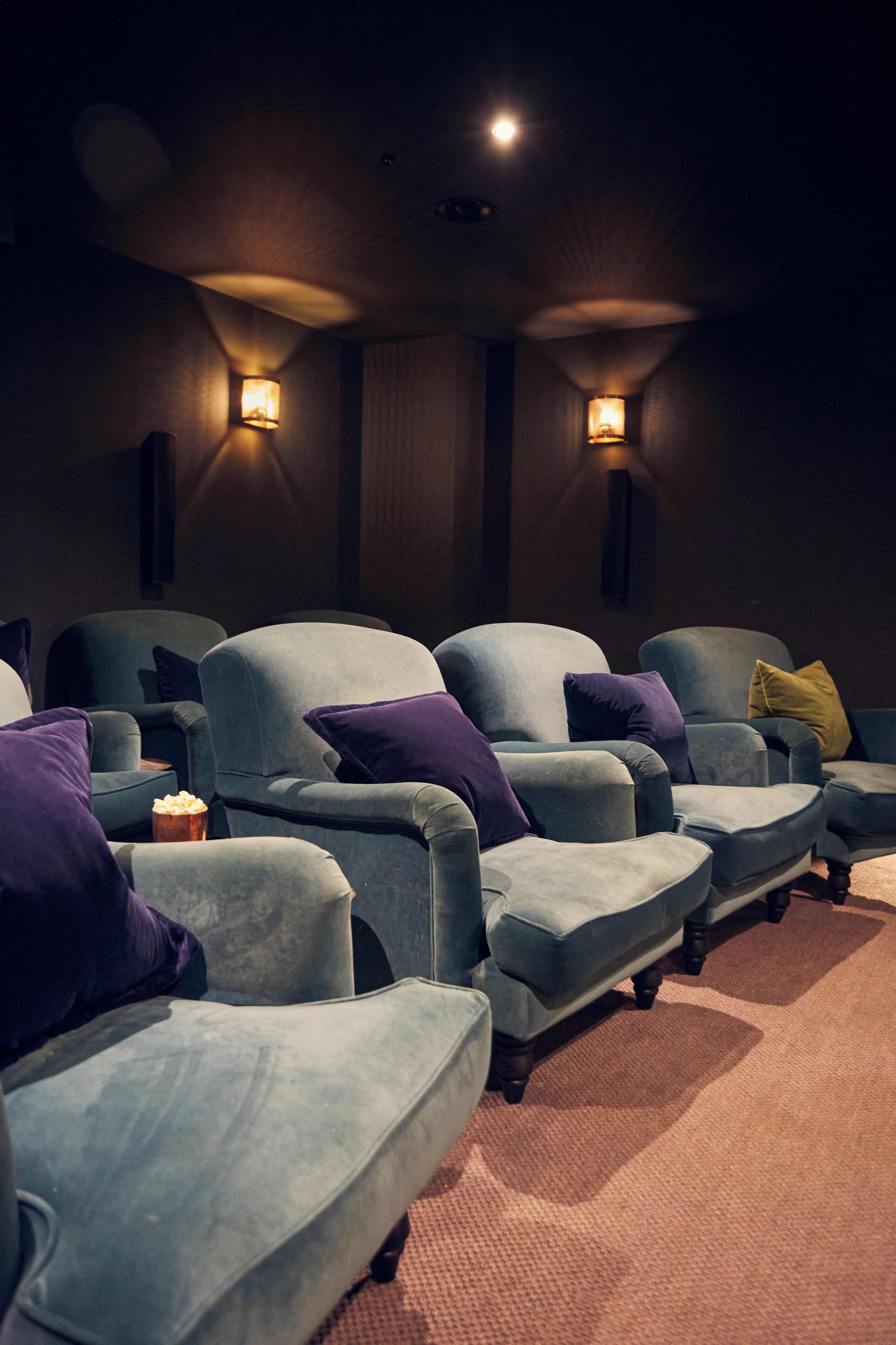 A row of blue armchairs with cushions, a popcorn bucket, and wall sconces in a dimly lit room.