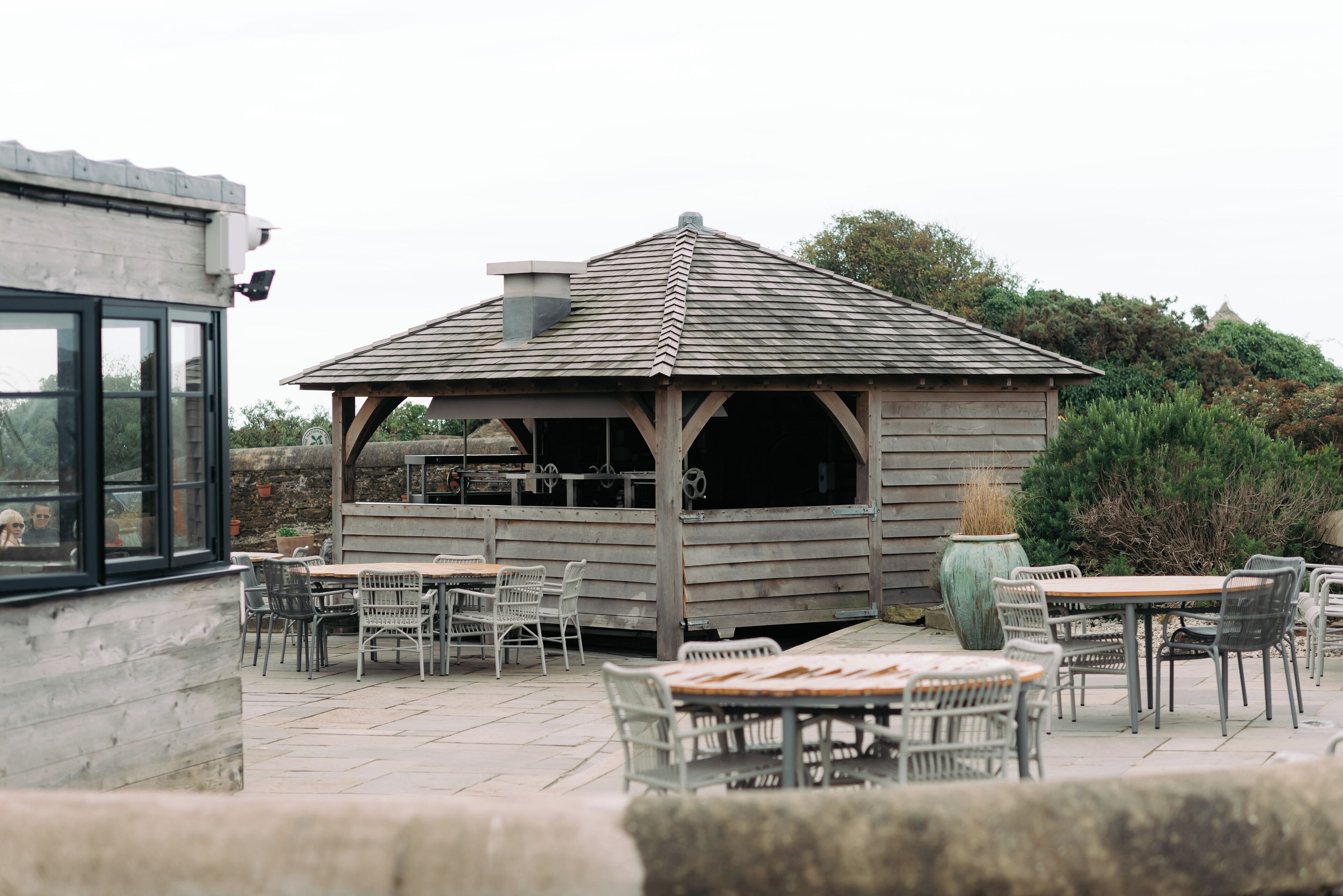Un espace extérieur aménagé en terrasse, avec des tables et des chaises en bois, une pergola et un mur en pierre.