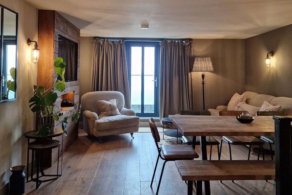 A living room with a sofa, armchair, dining table, and chairs. There is a large window with curtains, a plant, and wall-mounted lighting.