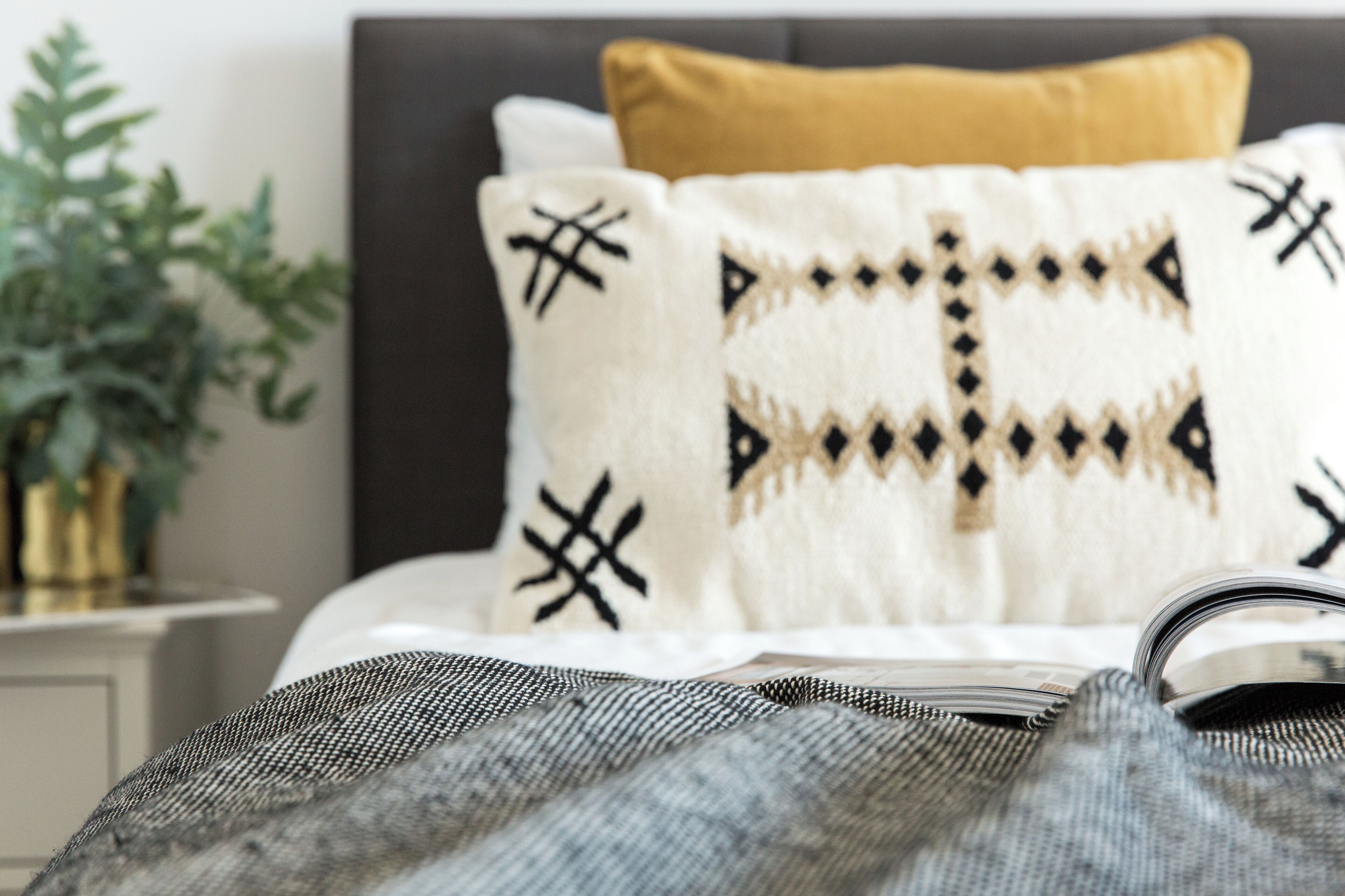 A bed with a patterned pillow, a book, and a plant in the background.