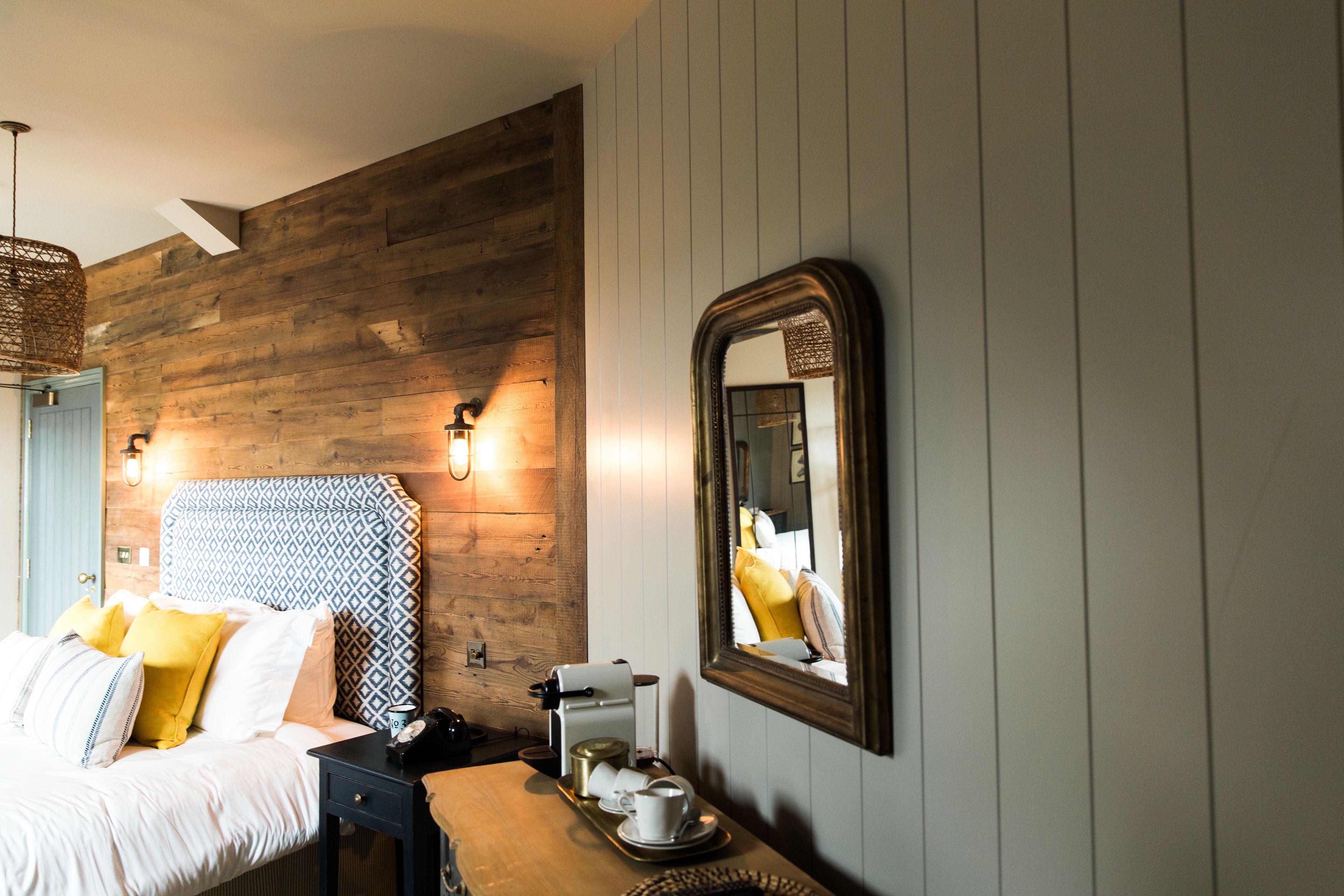 Ein Hotelzimmer mit einem Holz-Kopfteil, einem Bett mit weißer Bettwäsche und gelben Kissen, einem Nachttisch mit einer Kaffeemaschine und einem wandmontierten Spiegel.