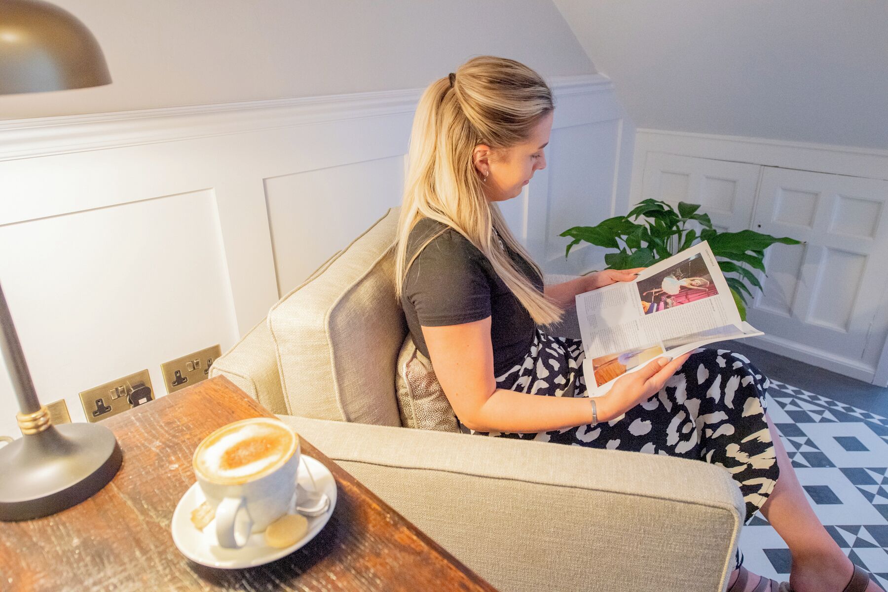 A person sitting on a sofa with a cup of coffee on a table beside them, reading a magazine.