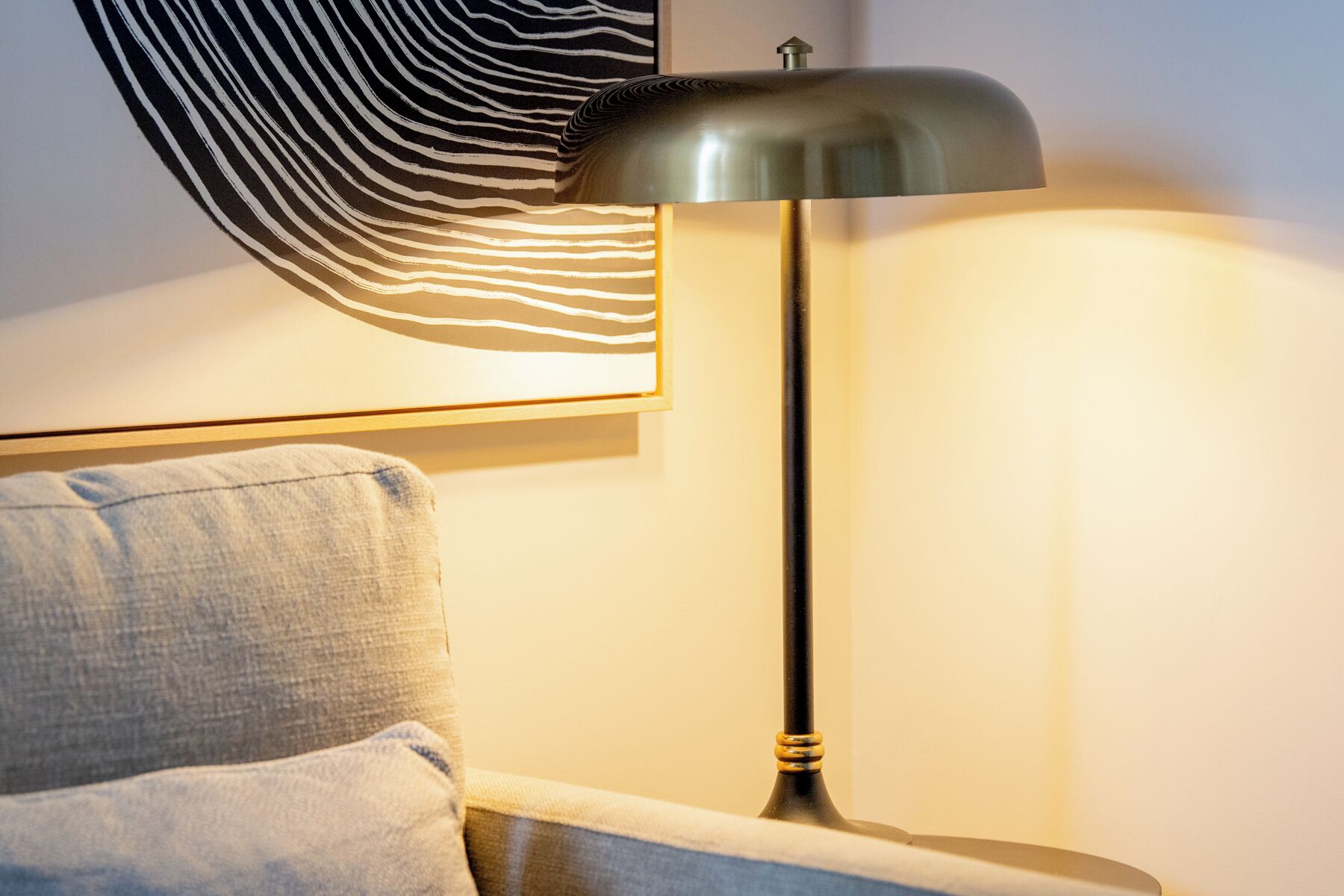 Une lampe moderne avec un abat-jour métallique, un élément décoratif mural texturé et un canapé de couleur claire.