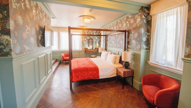 Una cama con dosel con un edredón rojo, un sillón rojo y una mesita de noche de madera con una lámpara.