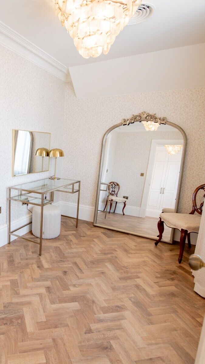 A room with a herringbone wooden floor, a glass desk with a gold frame, a large mirror, and a chandelier.