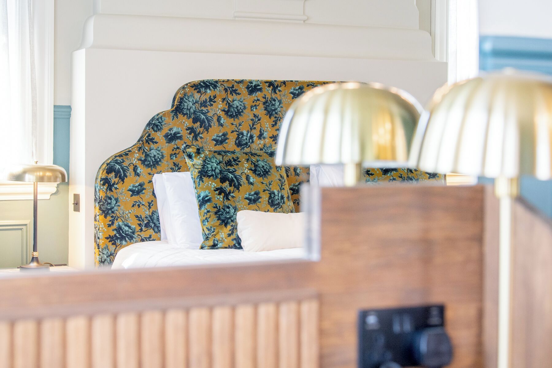 A hotel room with a floral-patterned headboard, a wooden bedside table, and two lamps.