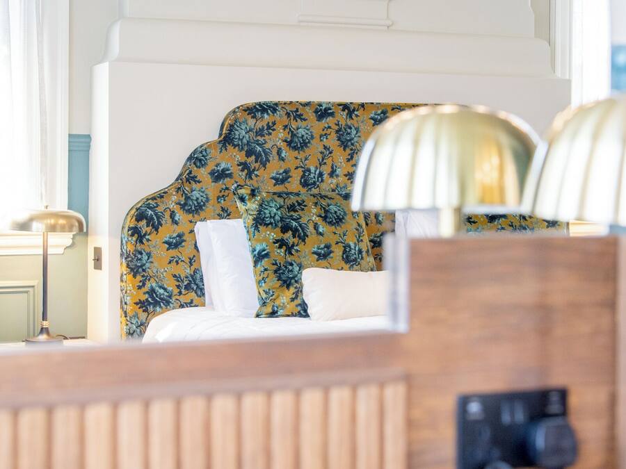 A hotel room with a floral-patterned headboard, a wooden bedside table, and two lamps.