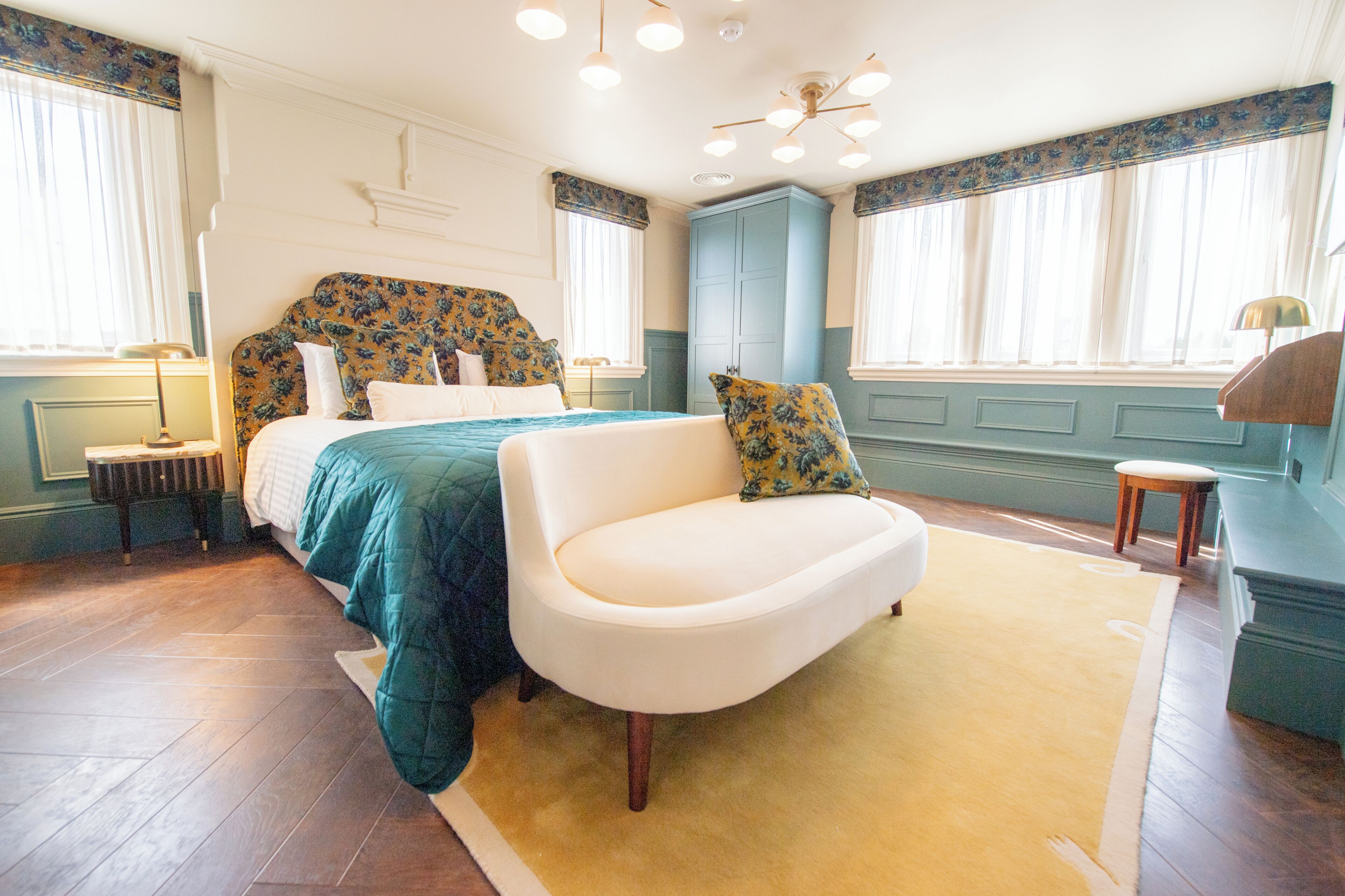 A bedroom with a bed, a white armchair, a nightstand, a lamp, a window with curtains, and a wardrobe.
