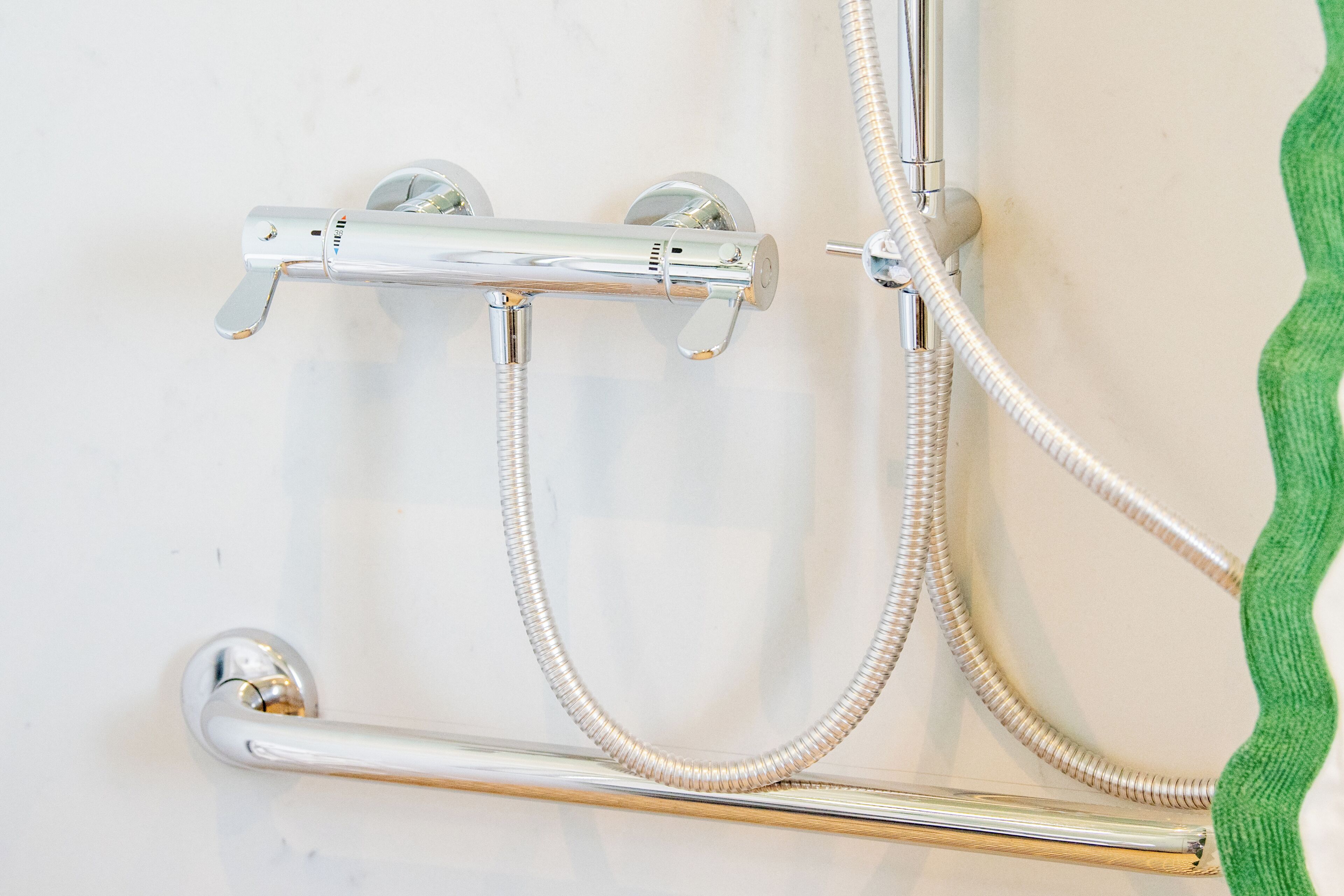 A shower with a chrome faucet and hose, a grab bar, and a green towel on a white wall.