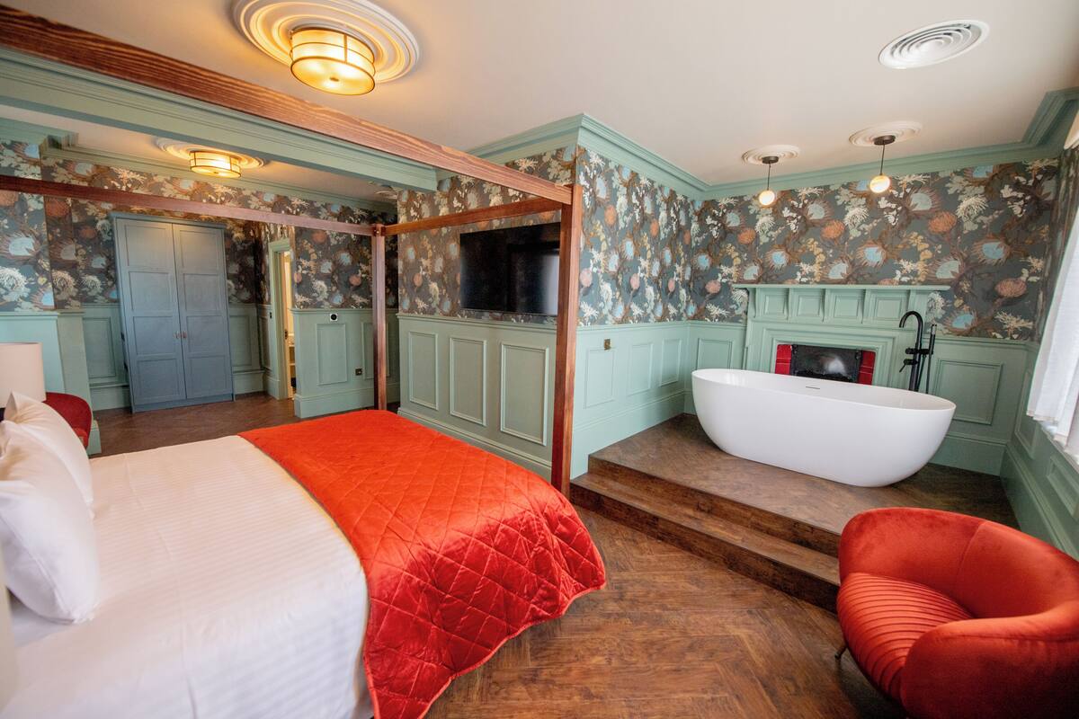 Ein Schlafzimmer mit einem Himmelbett, einem roten Sessel, einer weißen Badewanne und Holzboden.