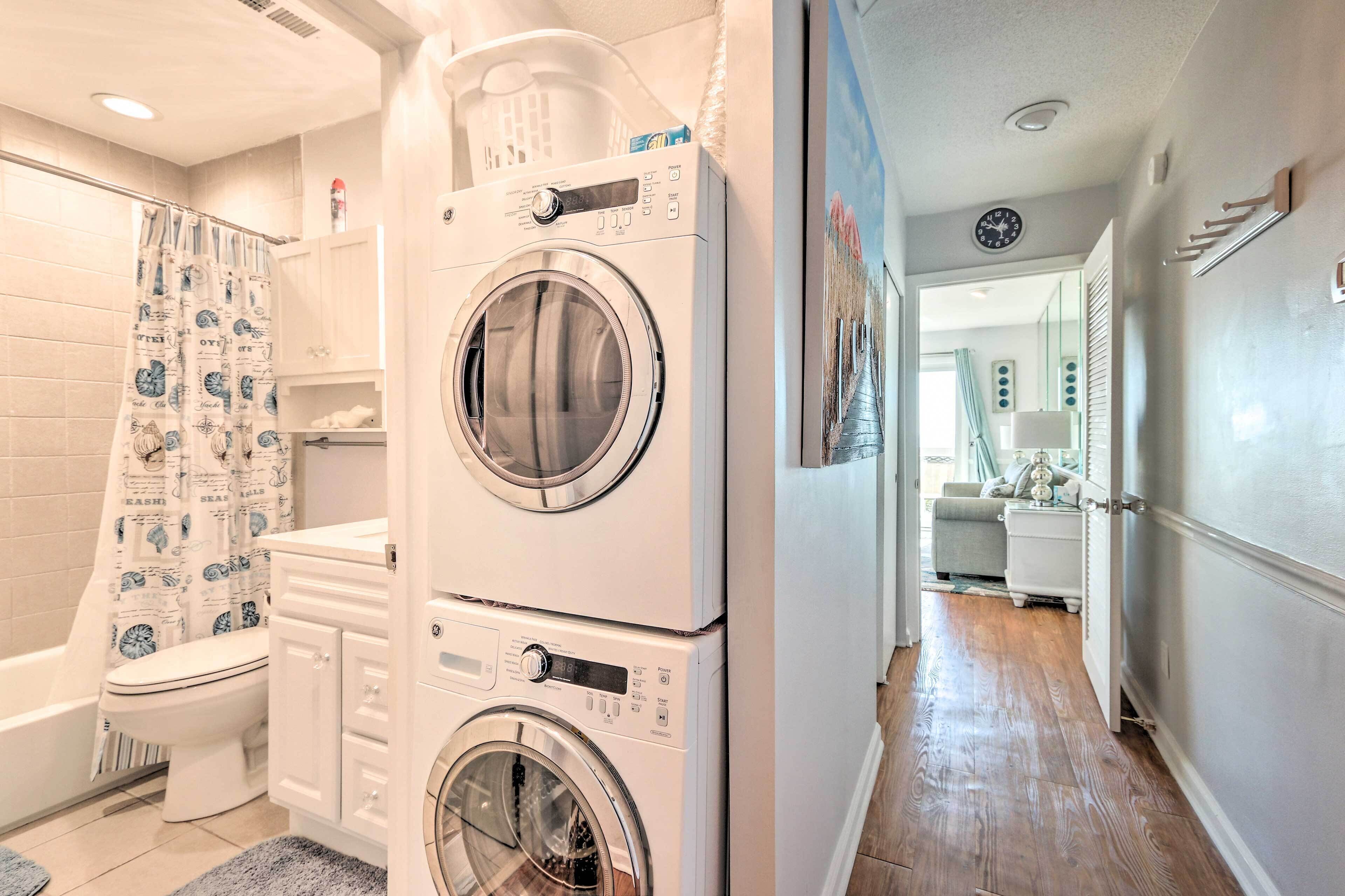 Un lavadero con lavadoras y secadoras apiladas, una ducha con cortina estampada y un pasillo que conduce a una sala de estar.