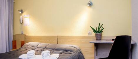 Habitación de hotel con cama, escritorio, silla, planta en maceta y teléfono.