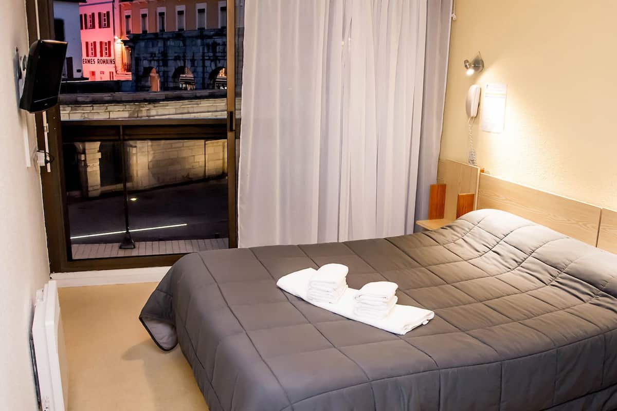 A hotel room with a bed, a window with curtains, a nightstand with a lamp, and a view of a building with a sign reading "ERMES ROMAINS".