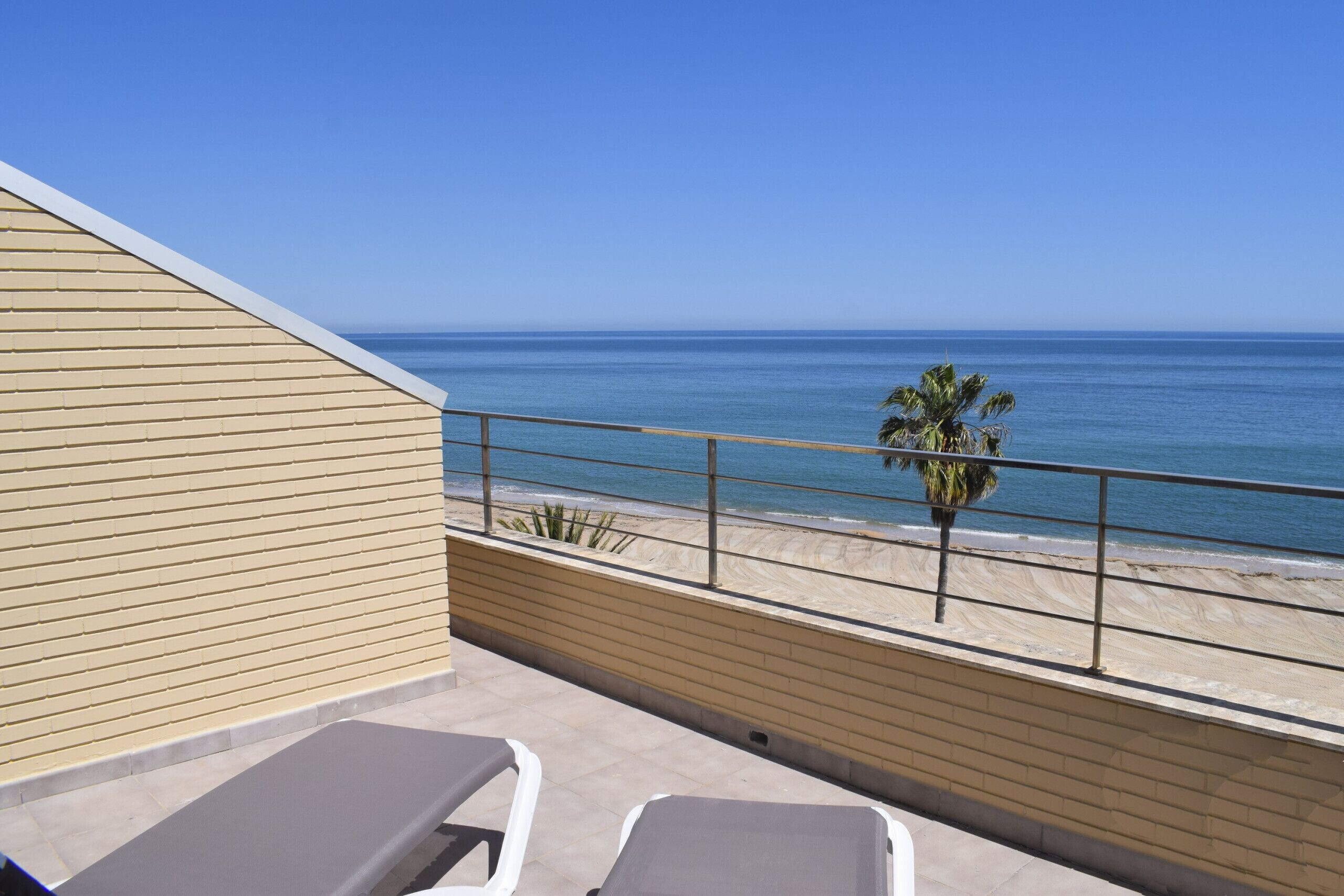 A balcony with two sun loungers, a view of the beach, and a palm tree.