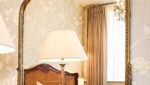 Um quarto com um espelho grande e trabalhado, uma mesa de cabeceira de madeira com livros, uma cama com encosto e uma janela com cortinas.