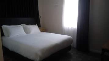 Quarto de hotel com uma cama grande, uma mesa de cabeceira de madeira, uma janela com cortinas e uma parede lisa.