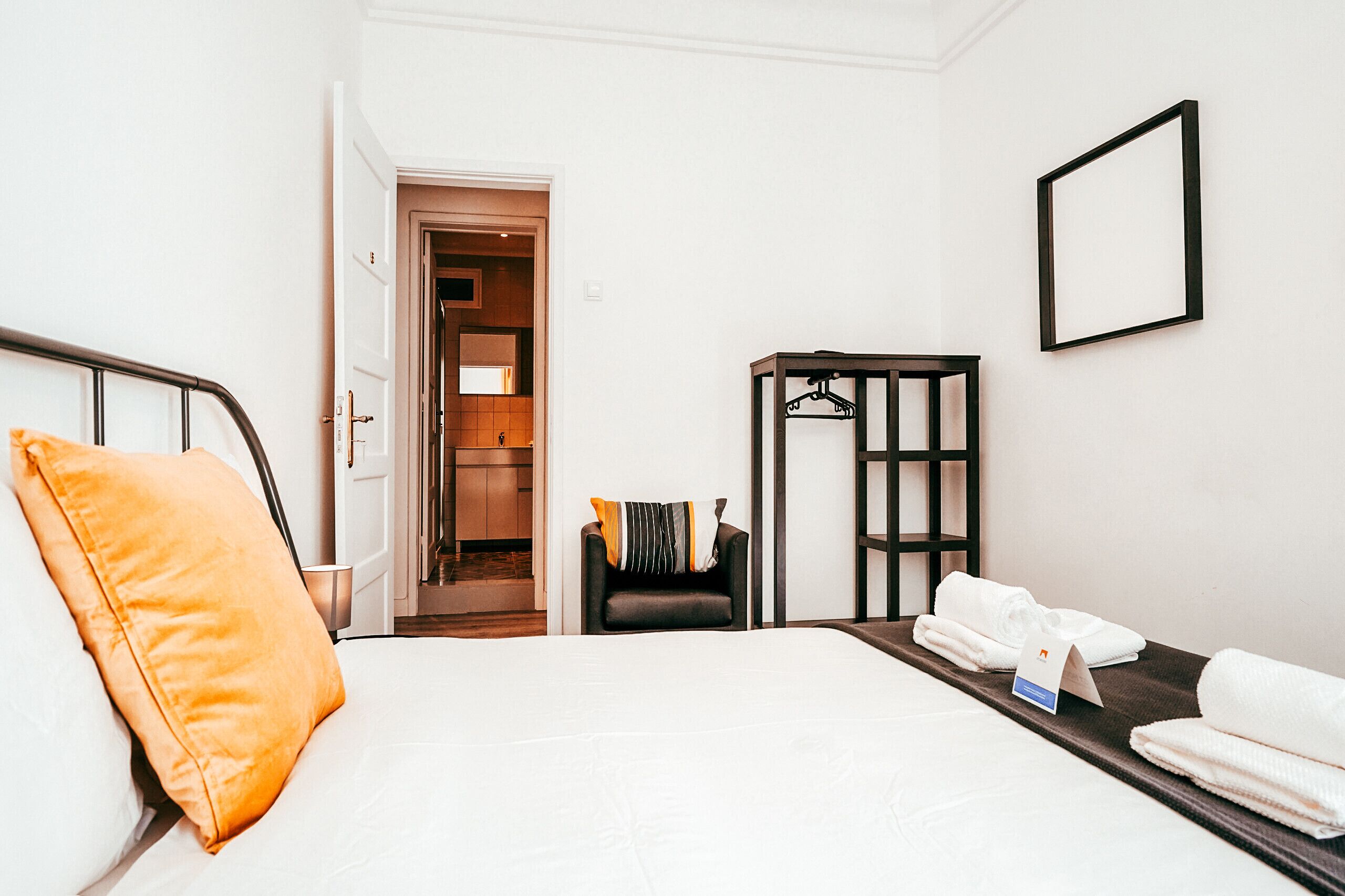 A hotel room with a bed, a chair, a coat rack, and a framed picture on the wall.