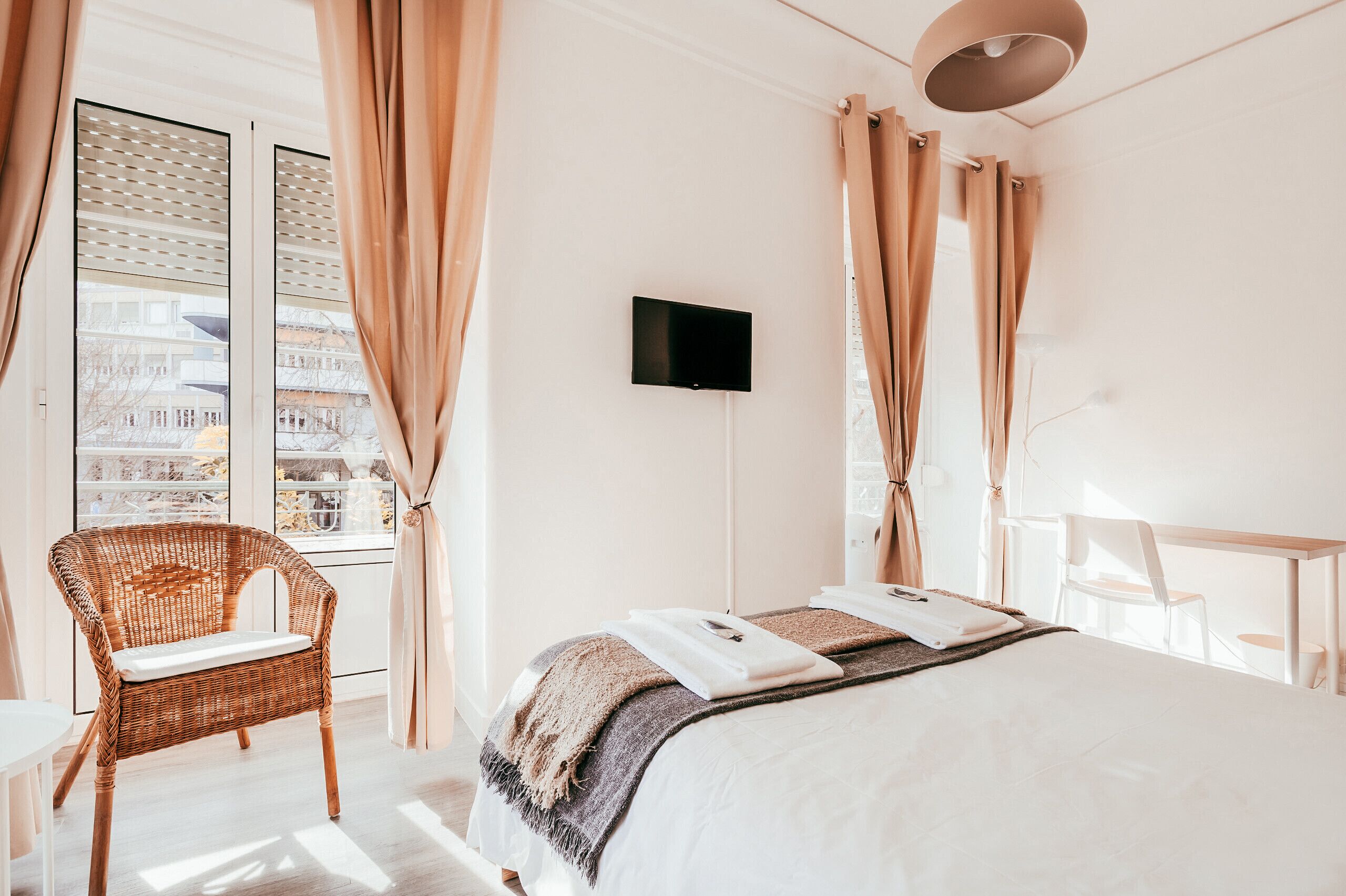 A bedroom with a bed, a wicker chair, a desk, and a TV on the wall.