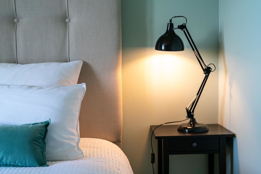 Una cama bien hecha con almohadas blancas y verde azulado, una mesita de noche pequeña con una lámpara de escritorio negra y un cabecero capitonado.