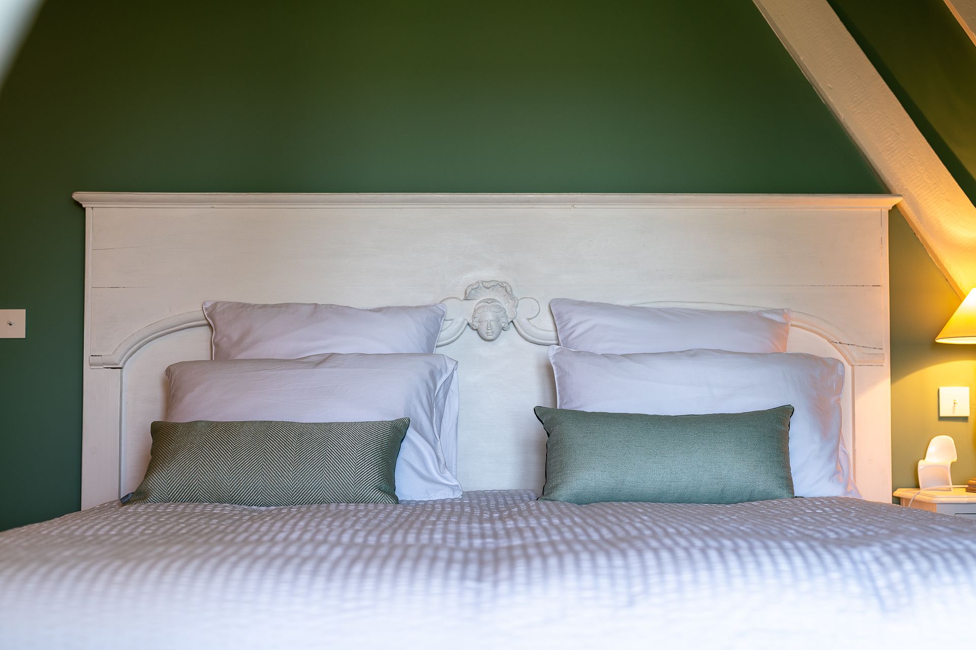 Un lit bien fait, avec une tête de lit blanche et deux oreillers, l’un à motifs et l’autre d’un vert uni, dans une pièce aux murs verts et au plafond en pente.