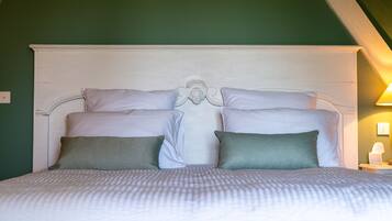 Un lit bien fait, avec une tête de lit blanche et deux oreillers, l’un à motifs et l’autre d’un vert uni, dans une pièce aux murs verts et au plafond en pente.