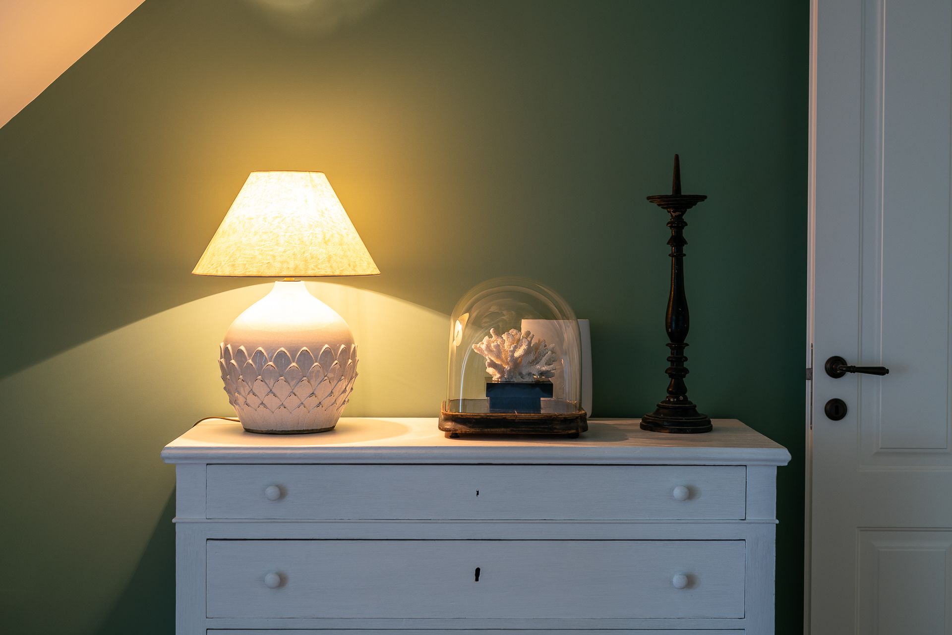 Une table de chevet blanche avec une lampe, un globe en verre contenant un corail, et un grand chandelier noir adossé à un mur vert.